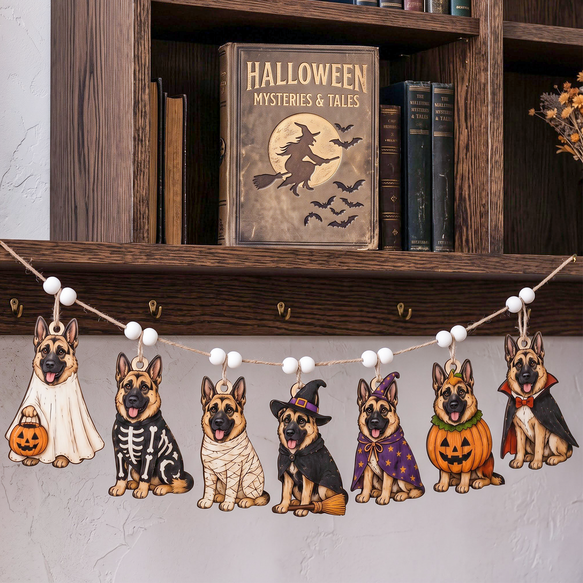 Halloween dog costume garland with six German Shepherd dog cutouts in different costumes hanging on a string with white beads, displayed in a home setting below a wooden bookshelf.