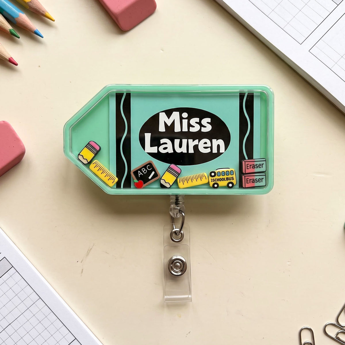 A mint green novelty desk accessory with a clear retractable badge reel, featuring a school-themed design with the text 'Miss Lauren' and small desk accessories like pencils, erasers, and paper clips on a beige desk surface.
