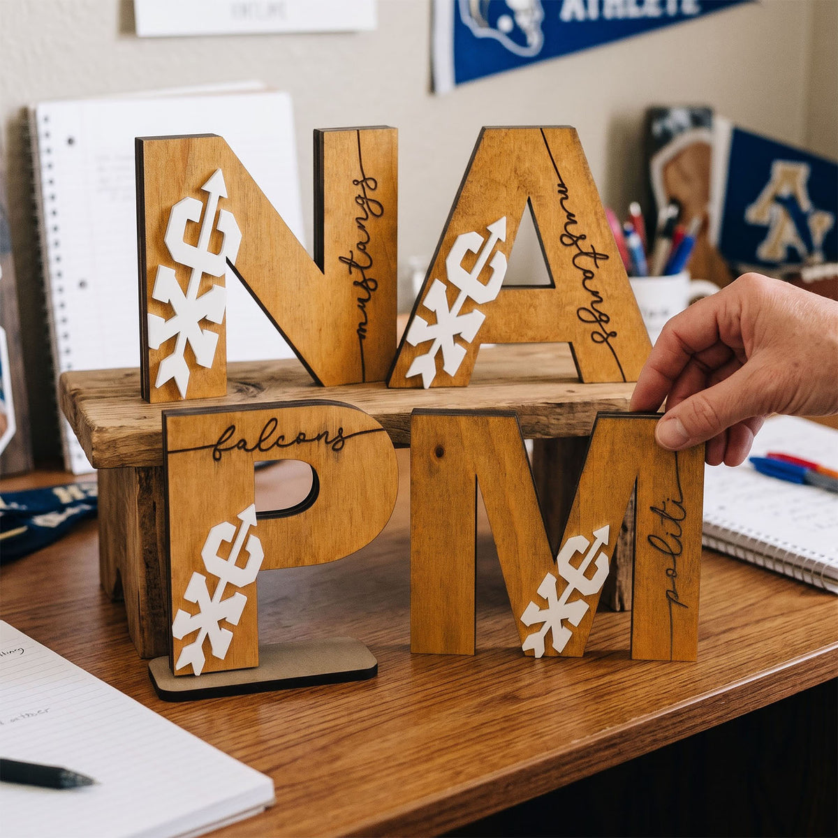 Wooden decorative letters spelling NAFLPM with white cutout graphics and black script text, displayed on a wooden stand on a desk with a hand holding one letter.