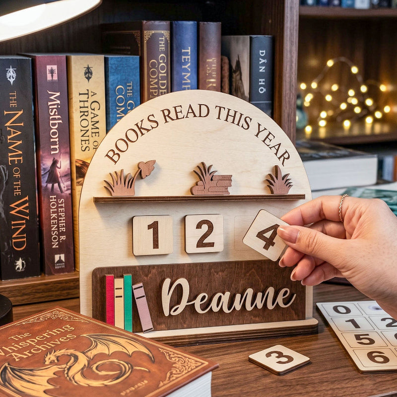 A wooden personalized bookshelf calendar stands on a desk in a cozy room setting. A hand is placing the number 4 on the year tiles.