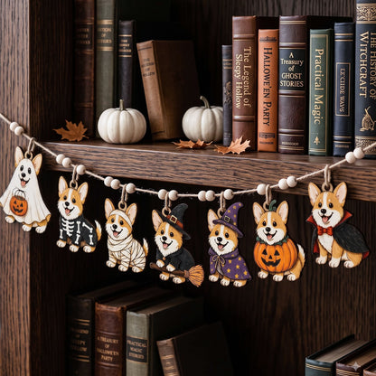 A Halloween-themed wooden dog garland draped across a dark wood bookshelf, featuring seven different corgi illustrations dressed in ghost, skeleton, bandage, witch, pumpkin, and vampire costumes, with small white beads on the string.