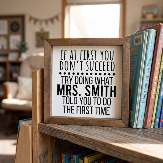 A square wooden framed sign with black text reading 'IF AT FIRST YOU DON'T SUCCEED TRY DOING WHAT MRS. SMITH TOLD YOU TO DO THE FIRST TIME' displayed on a wooden bookshelf next to books in a softly blurred indoor room.