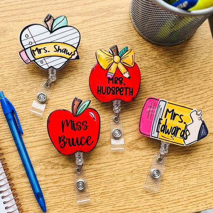Four personalized retractable badge reels shaped like an apple, a red apple, and a pencil, displayed on a light wood desk with a blue pen and a mesh cup in the background.