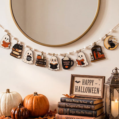 A Halloween-themed wooden garland with cute character cutouts hangs in front of a round gold-framed mirror. Below, pumpkins, a stack of books, a lantern, and a sign reading 'Happy Halloween' create a cozy seasonal display.