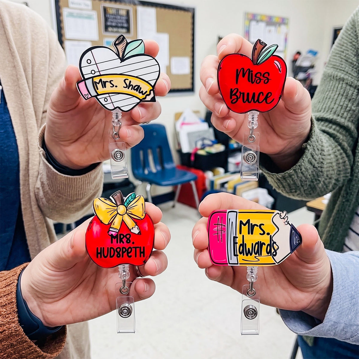 Four hands hold up custom school-themed bifold badge reels shaped like an apple, pencil, and two apples, each with a name printed on them. The background shows a classroom setting.