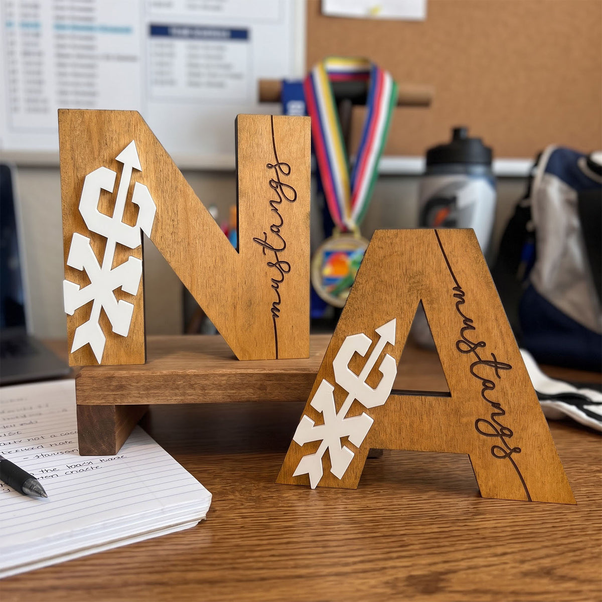 Three wooden decorative letters spelling "NA" displayed on a small wooden stand on a desk. The letters have white paper snowflake cutouts on the left and right sides and the word "mustang" written in black script down the center.