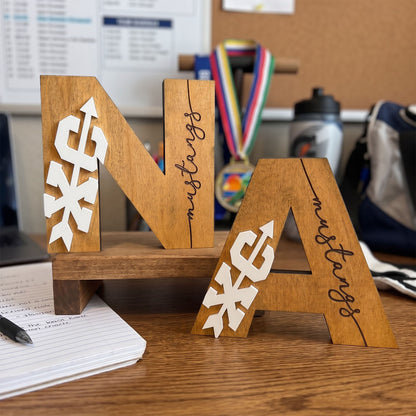 Three wooden decorative letters spelling "NA" displayed on a small wooden stand on a desk. The letters have white paper snowflake cutouts on the left and right sides and the word "mustang" written in black script down the center.