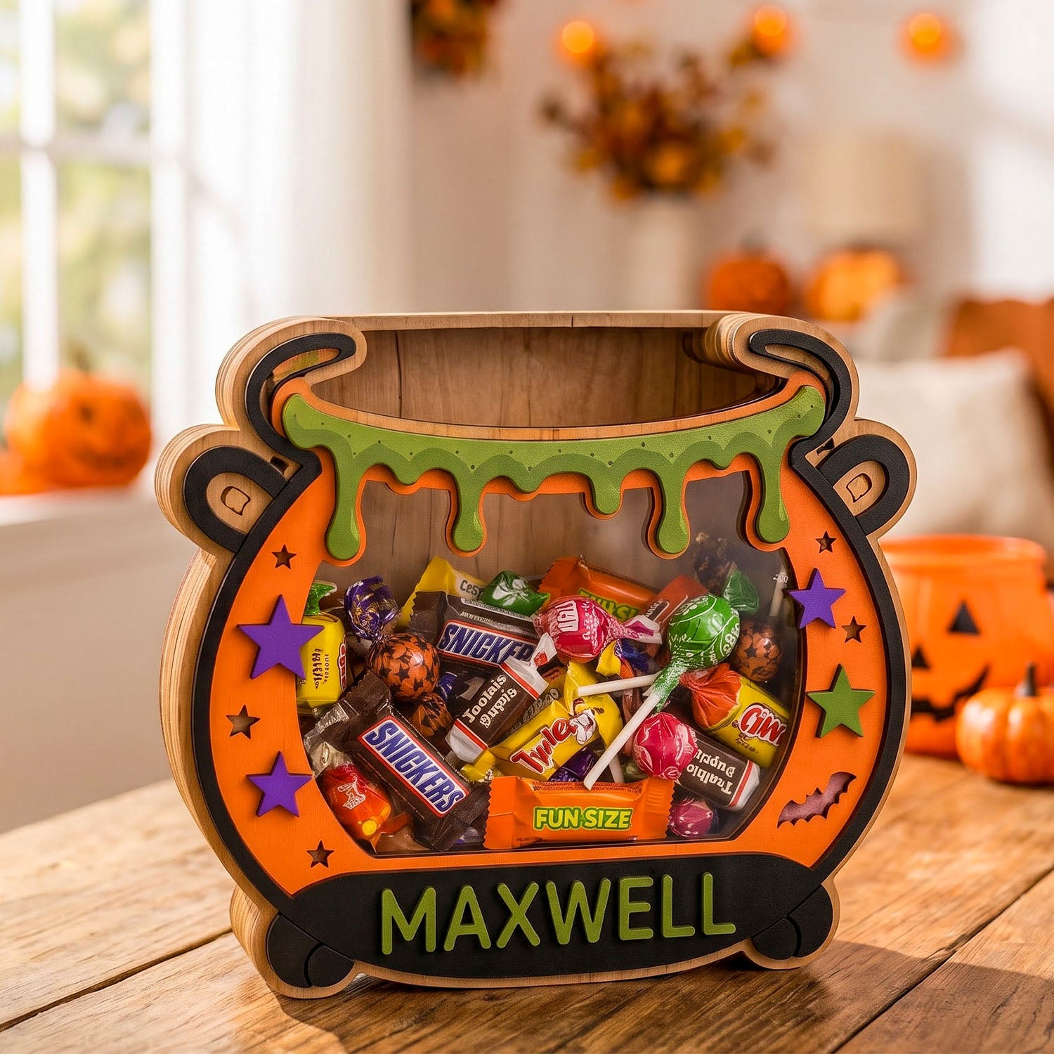 A Halloween-themed wooden cauldron-shaped candy box filled with assorted wrapped candies, placed on a wooden table in a decorated home setting. The front of the cauldron is personalized with the name 'MAXWELL' in green letters.