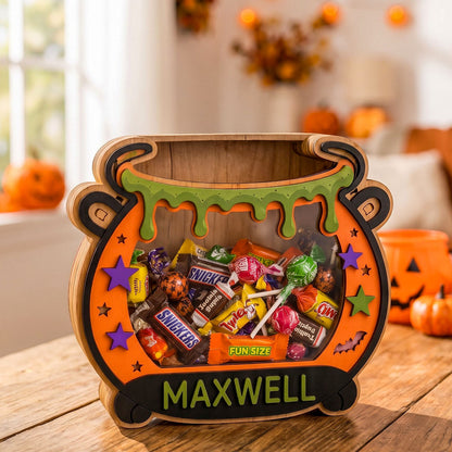 A Halloween-themed wooden cauldron-shaped candy box filled with assorted wrapped candies, placed on a wooden table in a decorated home setting. The front of the cauldron is personalized with the name 'MAXWELL' in green letters.