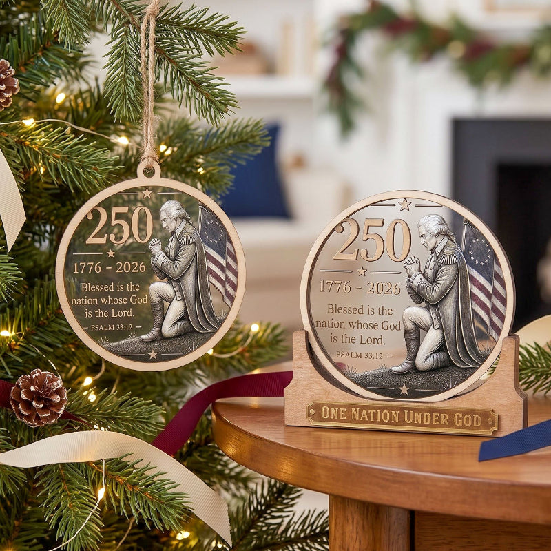 Two round wooden Christmas ornaments displayed on a table, one hanging on a decorated Christmas tree and one standing on a small base. The ornaments feature a silver-toned engraving of a kneeling praying man, the text '250 1776 - 2026 Blessed is the nation whose God is the Lord.