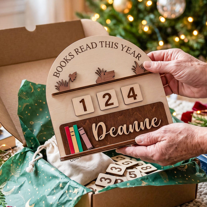 A wooden personalized book tracker plaque being held by hands over an open gift box. The plaque reads 'BOOKS READ THIS YEAR' with a year counter showing 1, 2, 4, and a custom name 'Deanne' on a dark wood panel.
