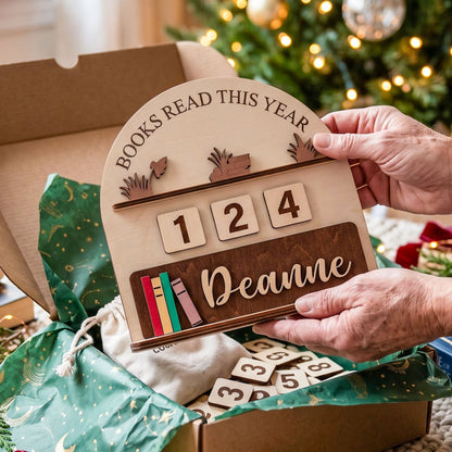 A wooden personalized book tracker plaque being held by hands over an open gift box. The plaque reads 'BOOKS READ THIS YEAR' with a year counter showing 1, 2, 4, and a custom name 'Deanne' on a dark wood panel.