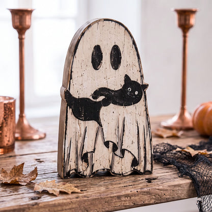 A Halloween decorative ghost figurine with a distressed off-white wood finish and a black cat printed across the front, displayed on a rustic wooden table with copper candlesticks, a pumpkin, and black netting in the background.