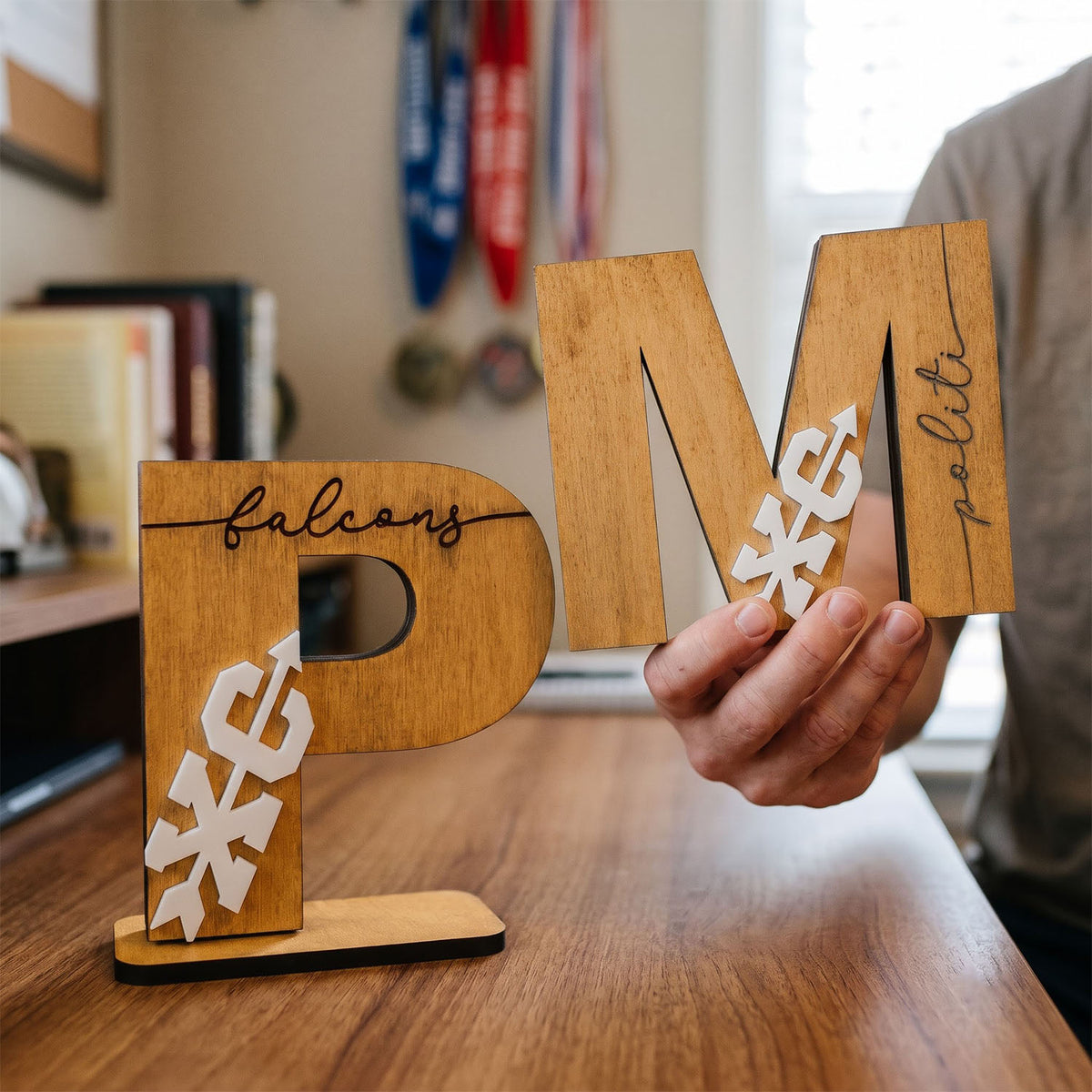 A person holds a large wooden letter 'M' with 'poli' written on it, while a freestanding wooden letter 'P' labeled 'falcon' sits on a table. The letters feature white eagle graphics and are shown in a home office setting.