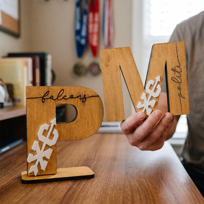 A person holds a large wooden letter 'M' with 'poli' written on it, while a freestanding wooden letter 'P' labeled 'falcon' sits on a table. The letters feature white eagle graphics and are shown in a home office setting.