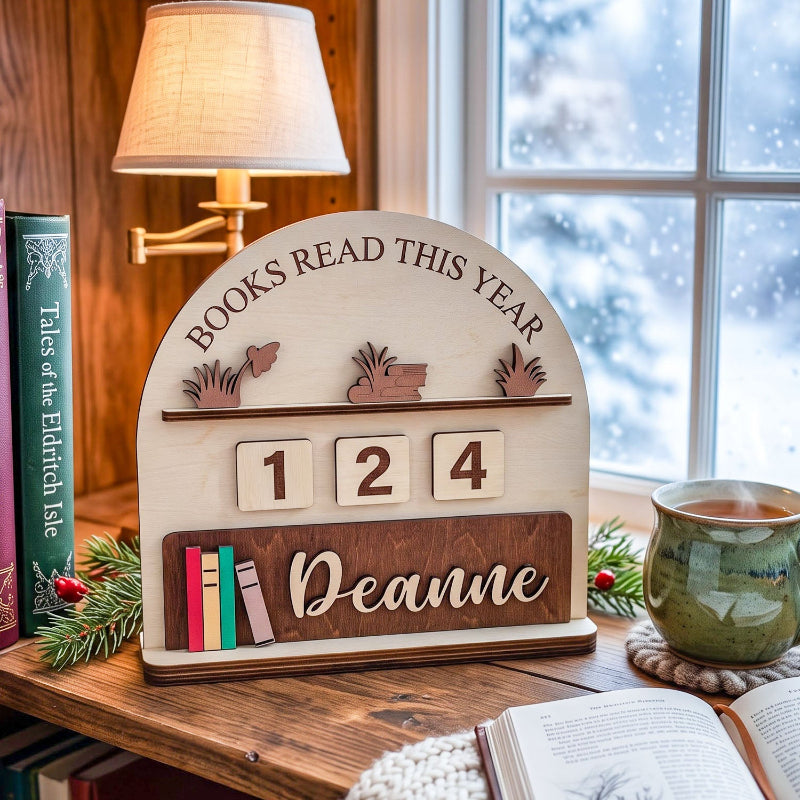 Personalized wooden bookshelf calendar sign on a rustic wooden table by a window, with a lamp, books, and a mug in a cozy room setting. The sign reads 'BOOKS READ THIS YEAR', has the numbers '1 2 4', and is personalized with the name 'Deanne'.