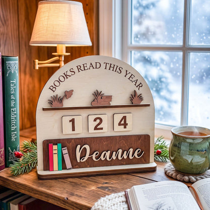 Personalized wooden bookshelf calendar sign on a rustic wooden table by a window, with a lamp, books, and a mug in a cozy room setting. The sign reads 'BOOKS READ THIS YEAR', has the numbers '1 2 4', and is personalized with the name 'Deanne'.