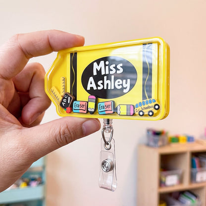 A hand holds a bright yellow pencil-shaped retractable badge reel with a clear plastic clip. The badge reel features school-themed graphics, including the text 'Miss Ashley', chalkboard markings, and small illustrations of erasers and a ruler.
