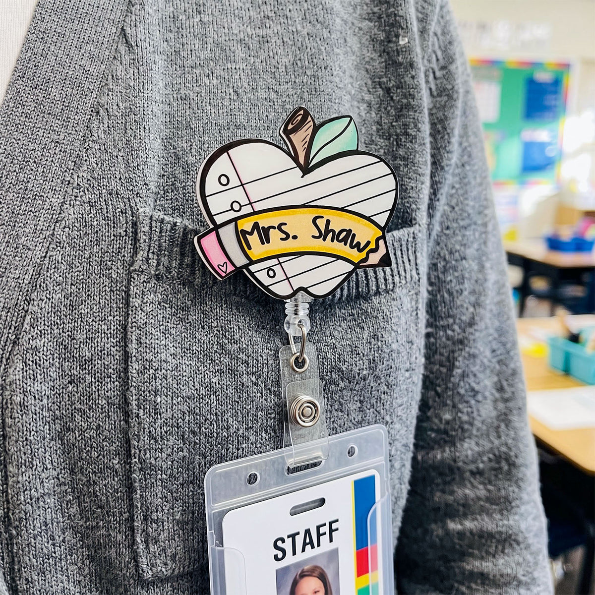 A heart-shaped enamel pin with a school notebook design, featuring the text 'Mrs. Shaws' on a yellow banner, pinned to a gray knit sweater.