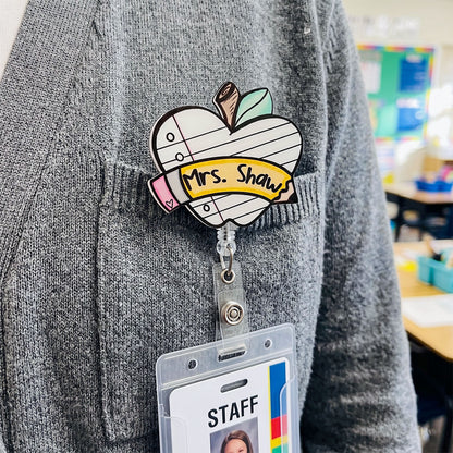A heart-shaped enamel pin with a school notebook design, featuring the text 'Mrs. Shaws' on a yellow banner, pinned to a gray knit sweater.