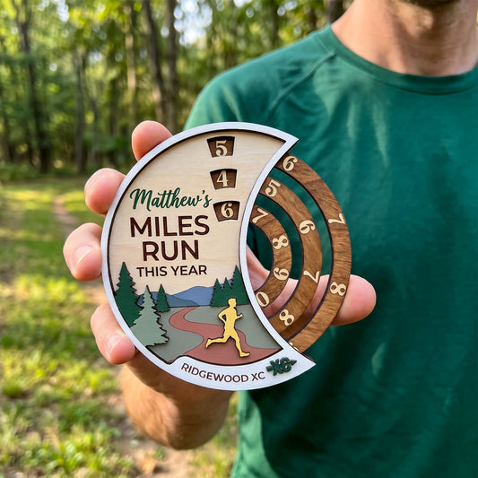 A person outdoors holding a personalized wooden running medallion that reads 'Matthew's MILES RUN THIS YEAR' with a small landscape illustration and numbered mile markers.