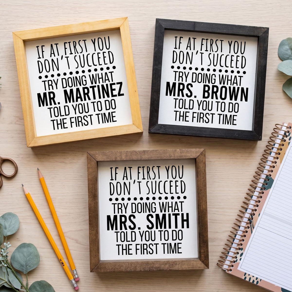 Three framed humorous signs with black text on white backgrounds, displayed on a light wood surface with decorative props including pencils, a notebook, and eucalyptus leaves.