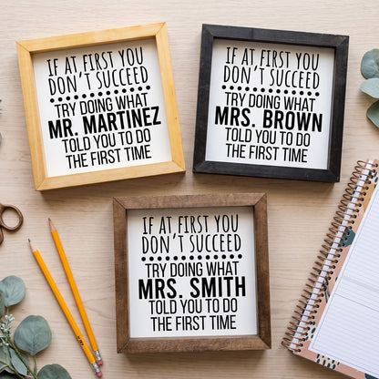 Three framed humorous signs with black text on white backgrounds, displayed on a light wood surface with decorative props including pencils, a notebook, and eucalyptus leaves.